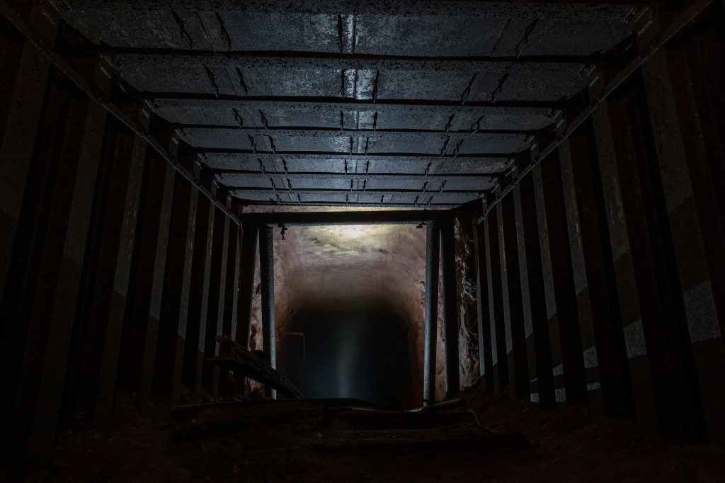 A dark, underground tunnel with metal supports and a faint light at the end, illuminating a water surface.