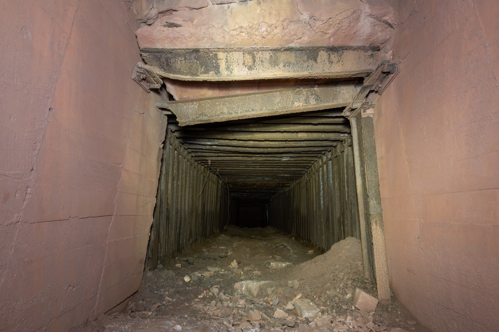 An underground tunnel with wooden supports, showing walls and a dusty floor partially covered with debris.
