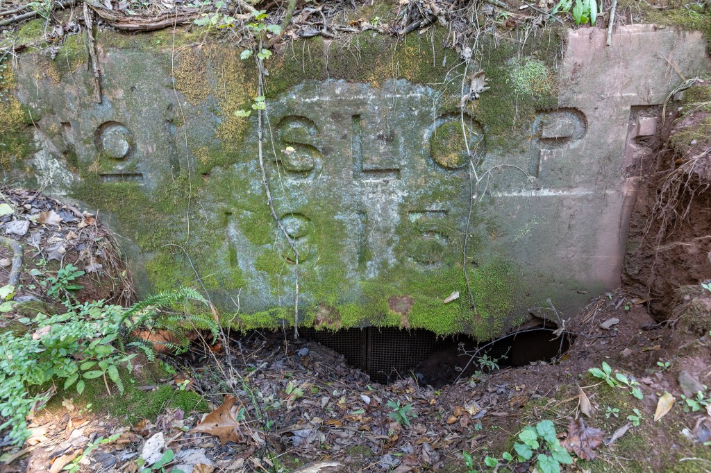 A weathered stone structure partially covered in moss and vegetation, featuring the engraved text 'NO SLOPE 1915', with an opening at the base revealing a dark interior.