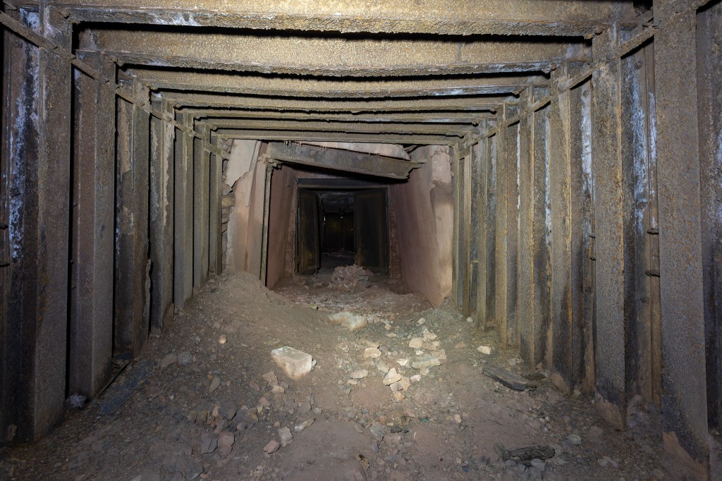An interior view of a dimly lit mining tunnel with metal beams supporting the walls and a pile of debris on the ground.